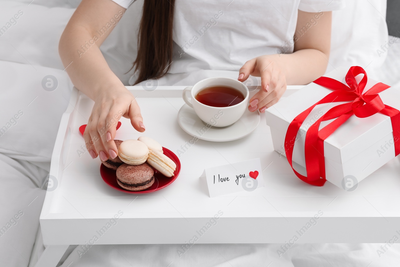 Photo of Tasty breakfast served in bed. Woman with tea, macarons, gift box and I Love You card at home, closeup