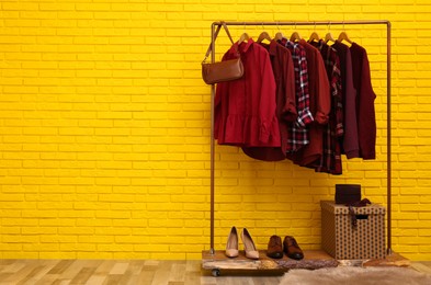 Rack with stylish clothes and bag near yellow brick wall indoors, space for text