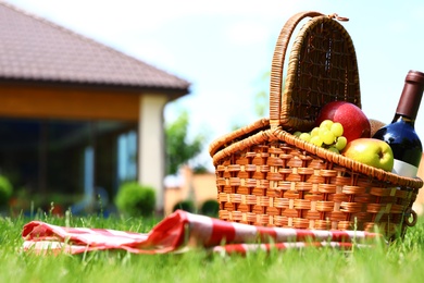 Picnic basket with fruits, bottle of wine and checkered blanket on green grass in garden. Space for text