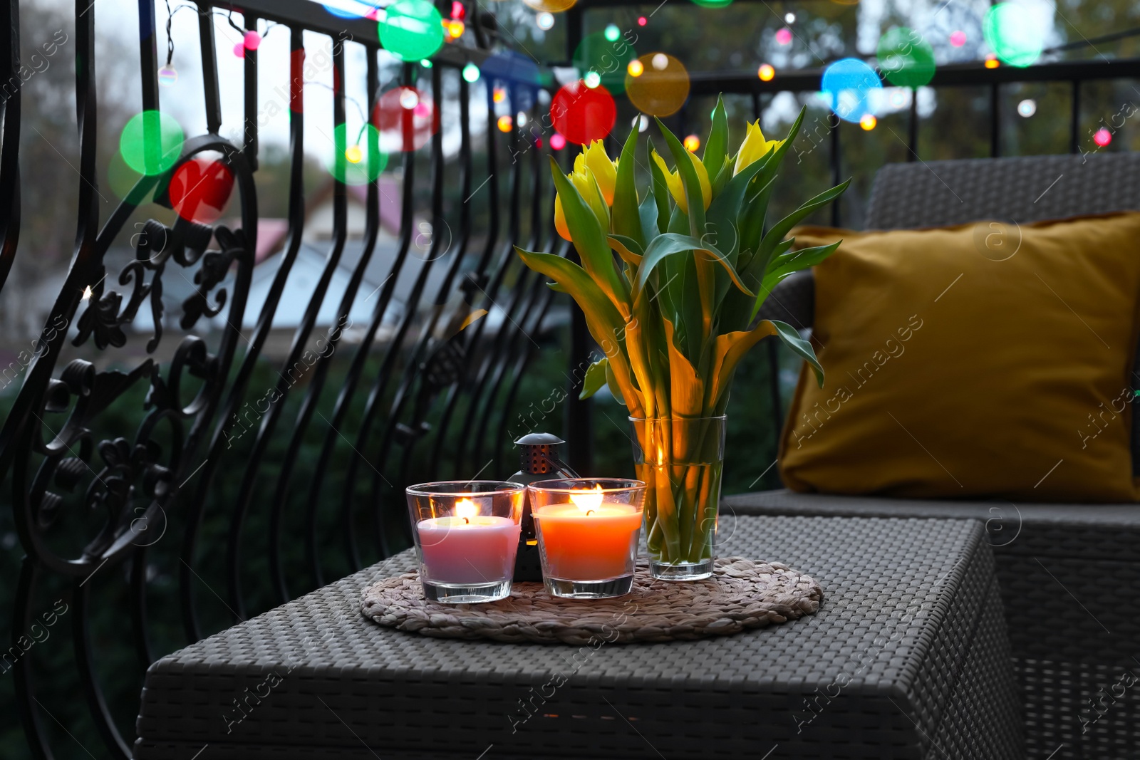 Photo of Soft pillows, blanket, burning candles and yellow tulips on rattan garden furniture in evening