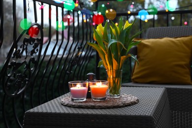 Soft pillows, blanket, burning candles and yellow tulips on rattan garden furniture in evening