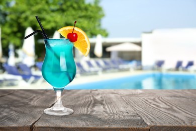 Image of Tasty Blue Lagoon cocktail on wooden table near outdoor swimming pool, space for text