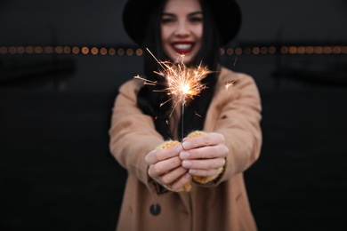 Photo of Woman in hat holding burning sparkler near building, focus on hands