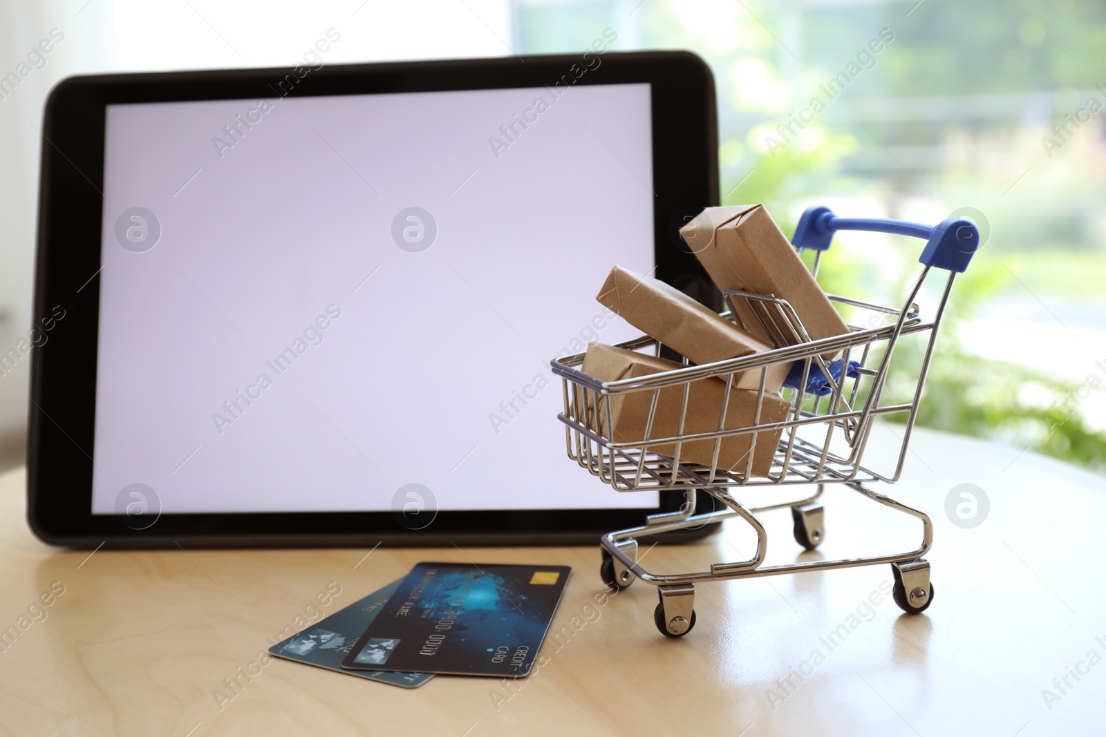 Photo of Internet shopping. Small cart with boxes and credit cards near modern tablet on table indoors