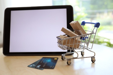 Internet shopping. Small cart with boxes and credit cards near modern tablet on table indoors