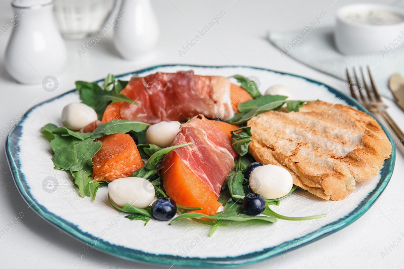 Photo of Fresh melon with prosciutto, mozzarella and arugula on white table