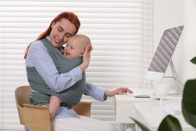 Photo of Mother holding her child in sling (baby carrier) while using computer at workplace