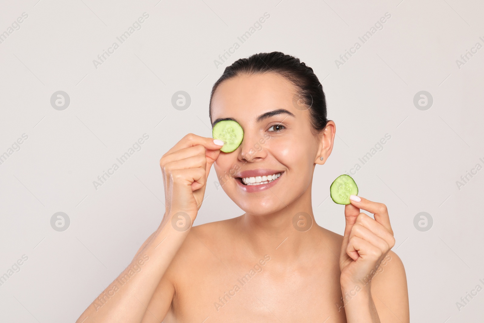 Photo of Beautiful young woman putting slices of cucumber on eyes against light grey background