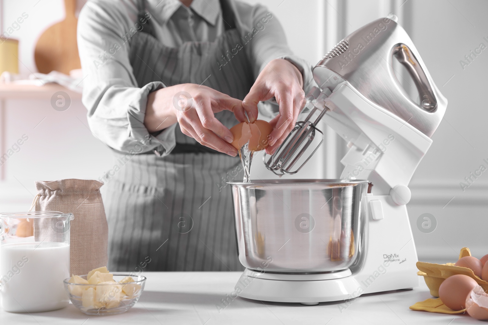 Photo of Woman adding egg into bowl of stand mixer while making dough at table indoors, closeup