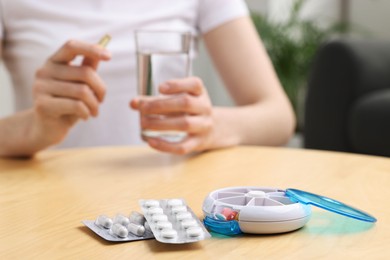 Woman with pills, organizer and glass of water at light wooden table, selective focus