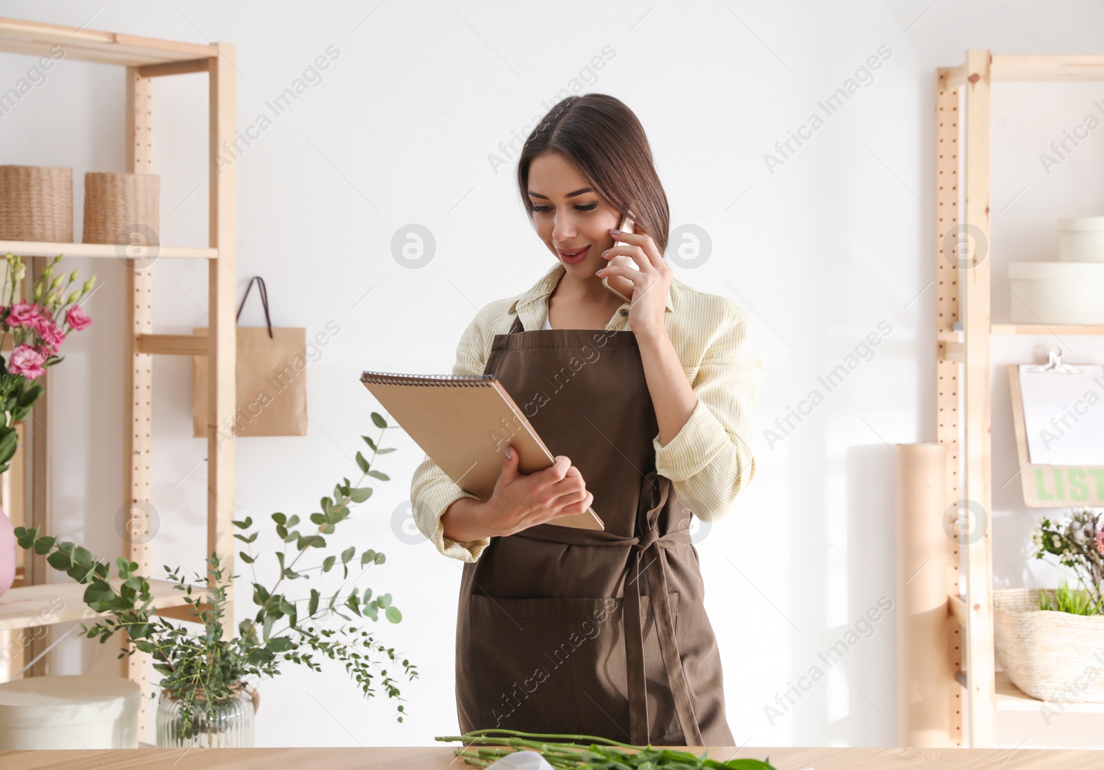 Photo of Florist with notebook talking on smartphone in workshop