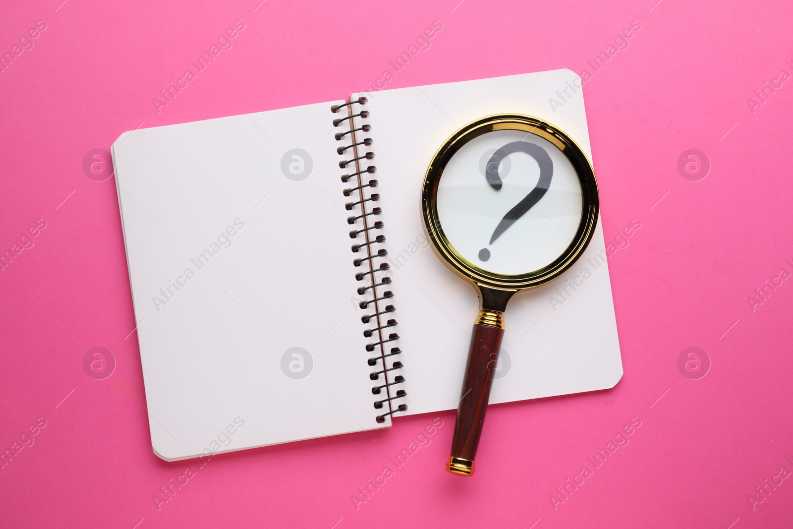 Photo of Magnifying glass over notebook with question mark on pink background, top view
