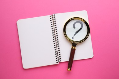 Magnifying glass over notebook with question mark on pink background, top view