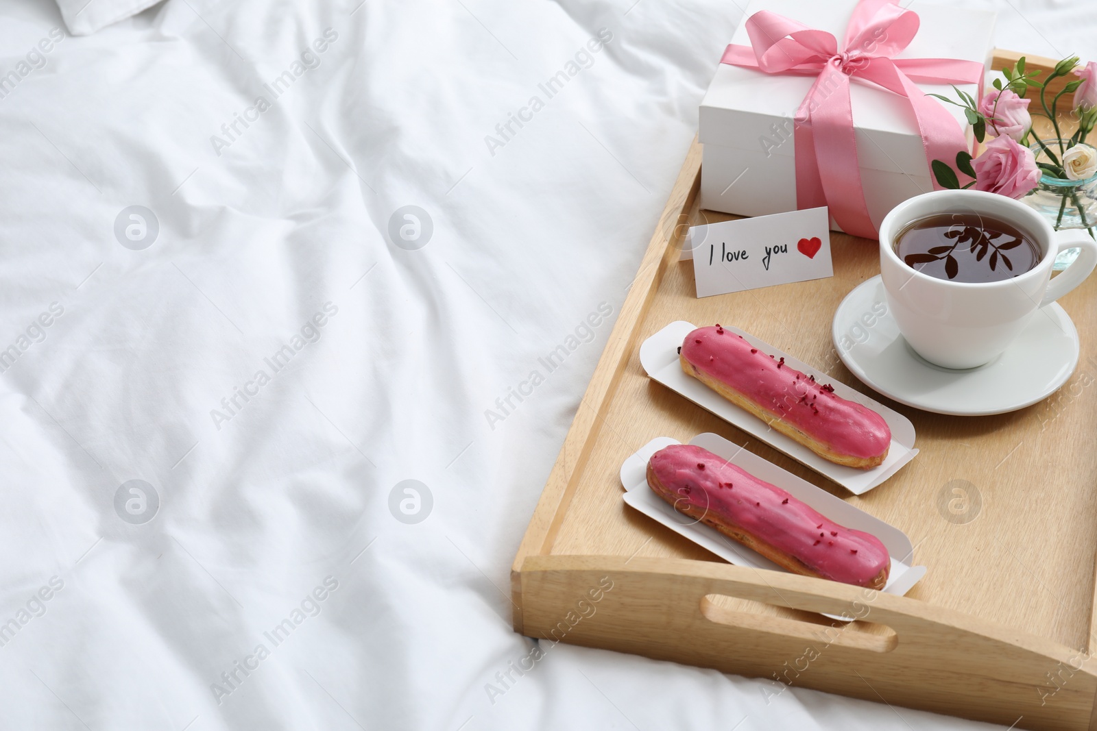 Photo of Tasty breakfast served in bed. Delicious eclairs, tea, gift box, flowers and card with phrase I Love You on tray, space for text