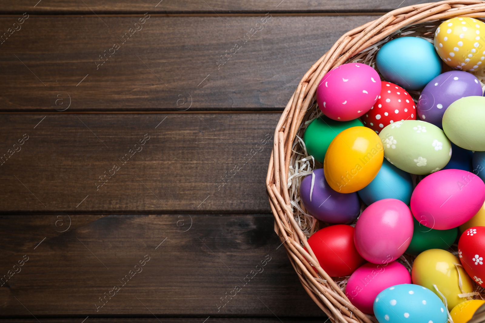 Photo of Colorful Easter eggs in wicker basket on wooden table, top view. Space for text