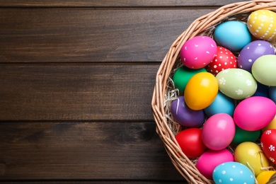 Photo of Colorful Easter eggs in wicker basket on wooden table, top view. Space for text