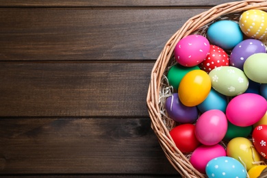 Colorful Easter eggs in wicker basket on wooden table, top view. Space for text