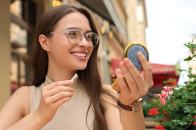 Beautiful young woman with cosmetic pocket mirror applying lipstick outdoors