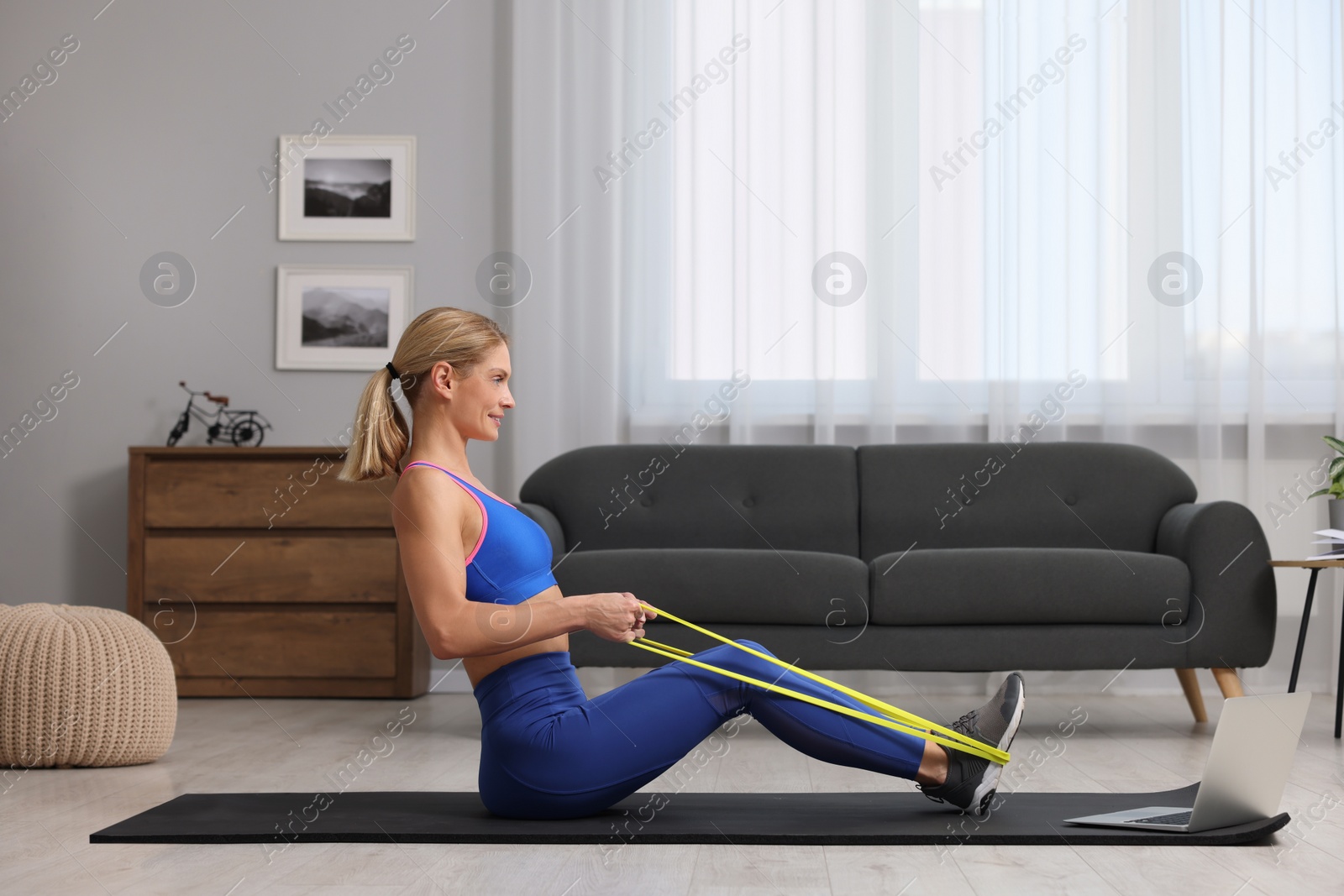Photo of Fit woman doing exercise with fitness elastic band near laptop on mat at home