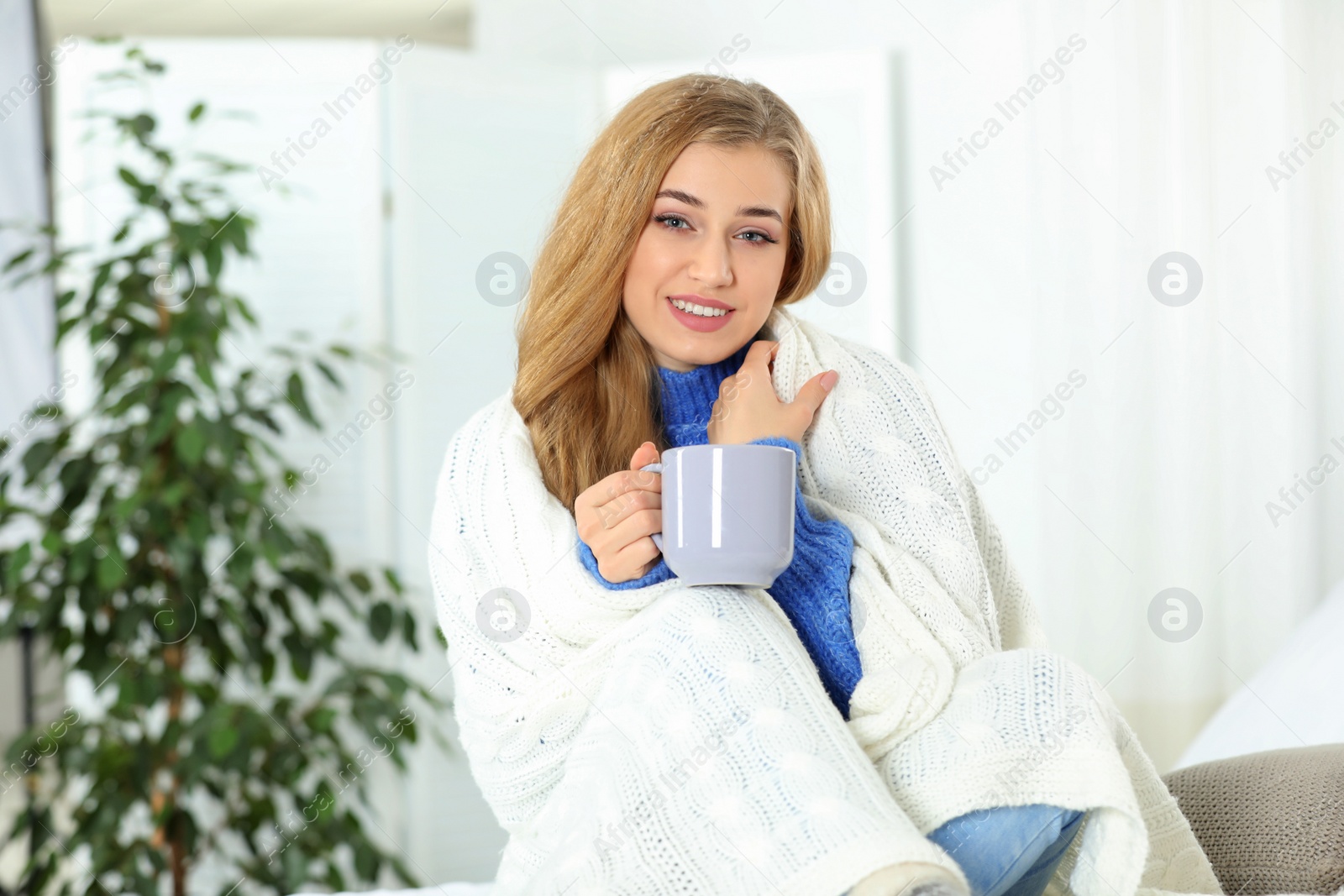 Photo of Attractive young woman in cozy warm sweater with cup of hot drink and plaid at home