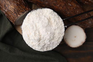 Photo of Scoop with cassava flour and root on wooden table, top view