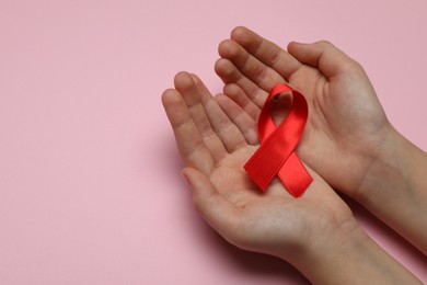 Little girl holding red ribbon on pink background, closeup with space for text. AIDS disease awareness