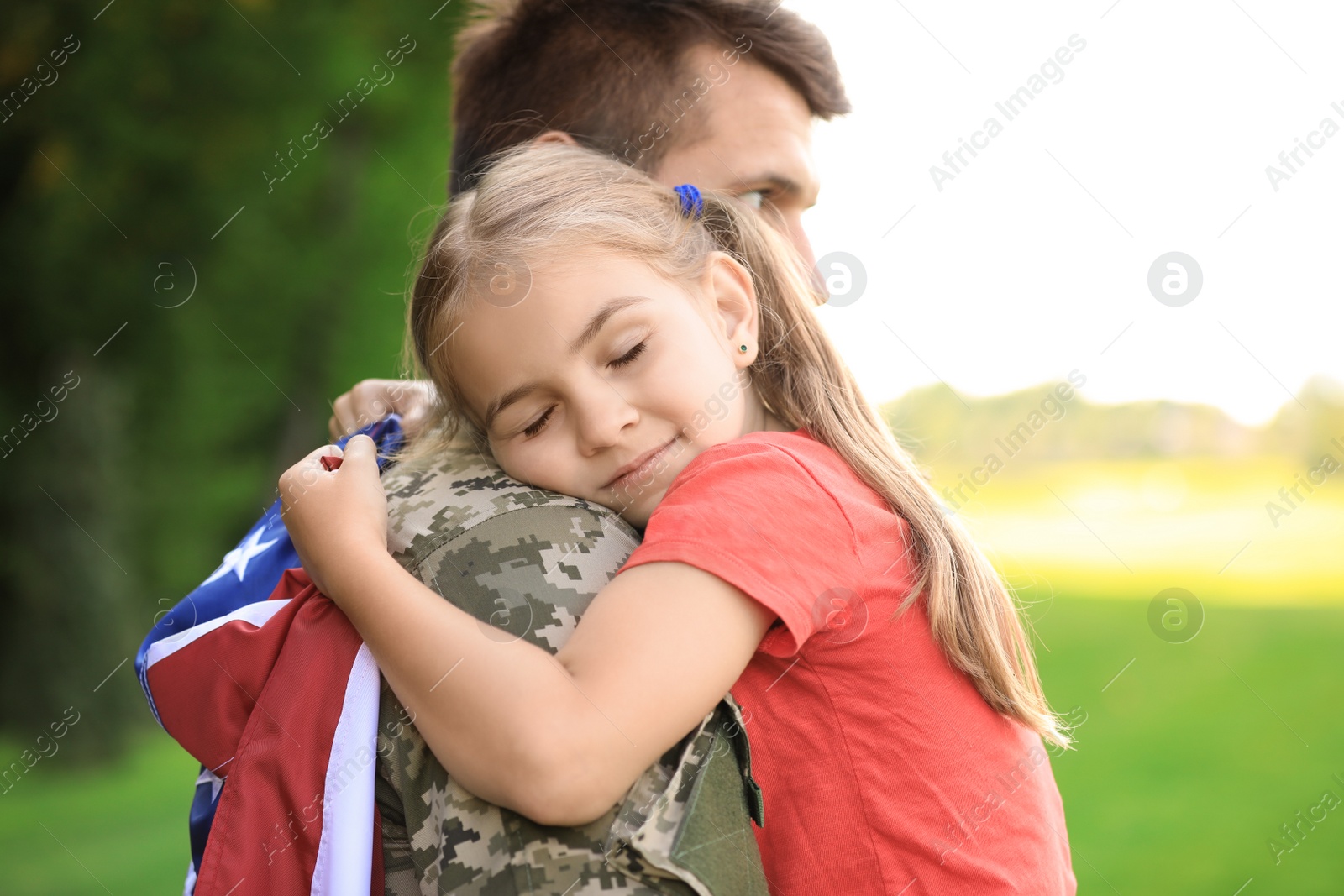 Photo of Father in military uniform with American flag hugging his little daughter at green park