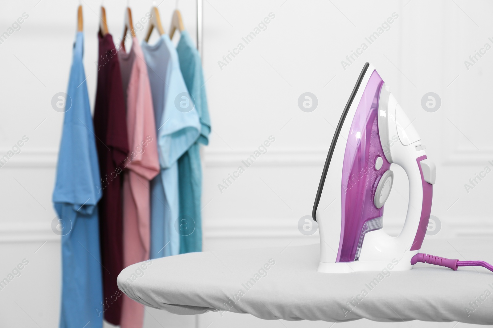 Photo of Modern iron on board near rack with clean clothes indoors. Space for text