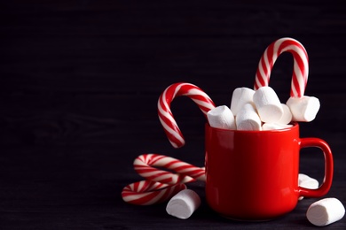 Photo of Cup of tasty cocoa with marshmallows and Christmas candy canes on black wooden table. Space for text