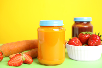 Jars with baby food and fresh ingredients on green table against yellow background