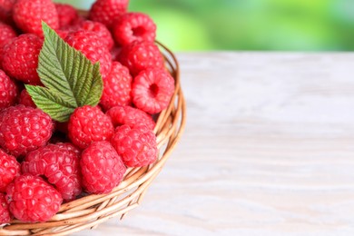 Tasty ripe raspberries and green leaf in wicker basket on white wooden table, closeup. Space for text
