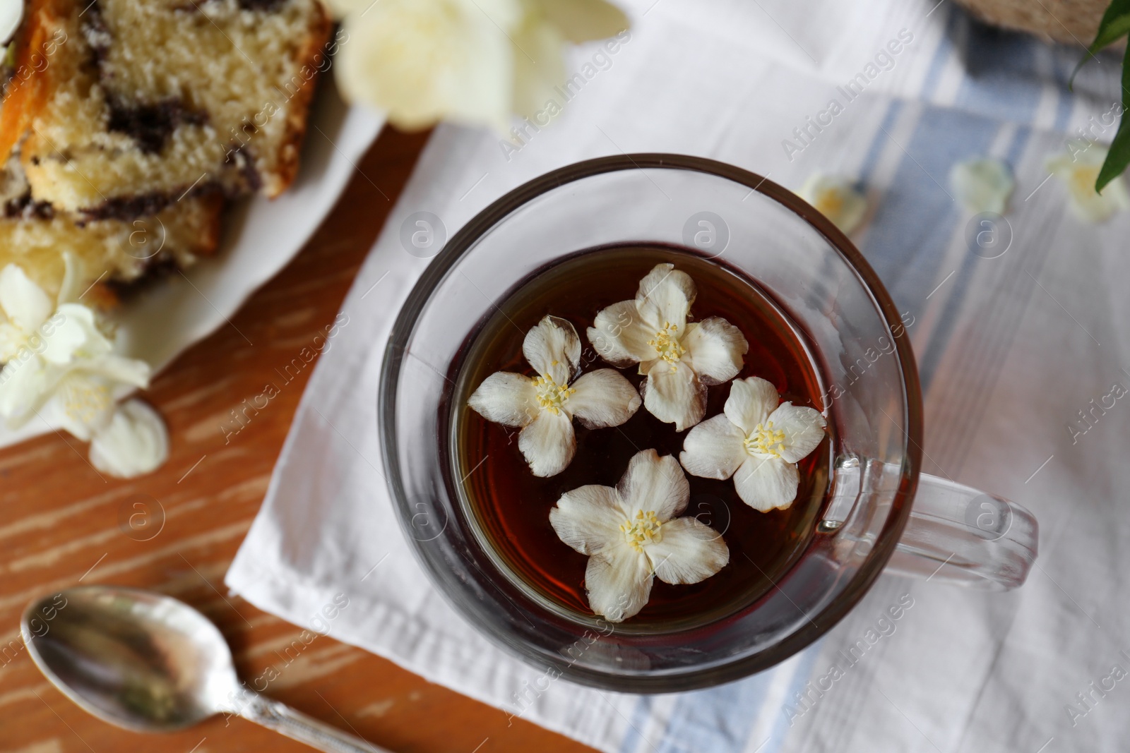 Photo of Glass cup of aromatic tea with beautiful jasmine flowers and dessert on wooden table, flat lay