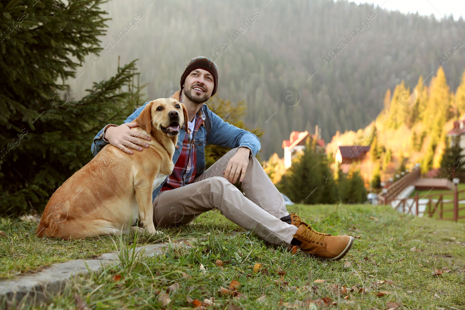 Photo of Happy man and adorable dog sitting on green grass in mountains. Traveling with pet