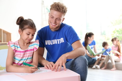 Photo of Little girl learning alphabet with volunteer at table indoors