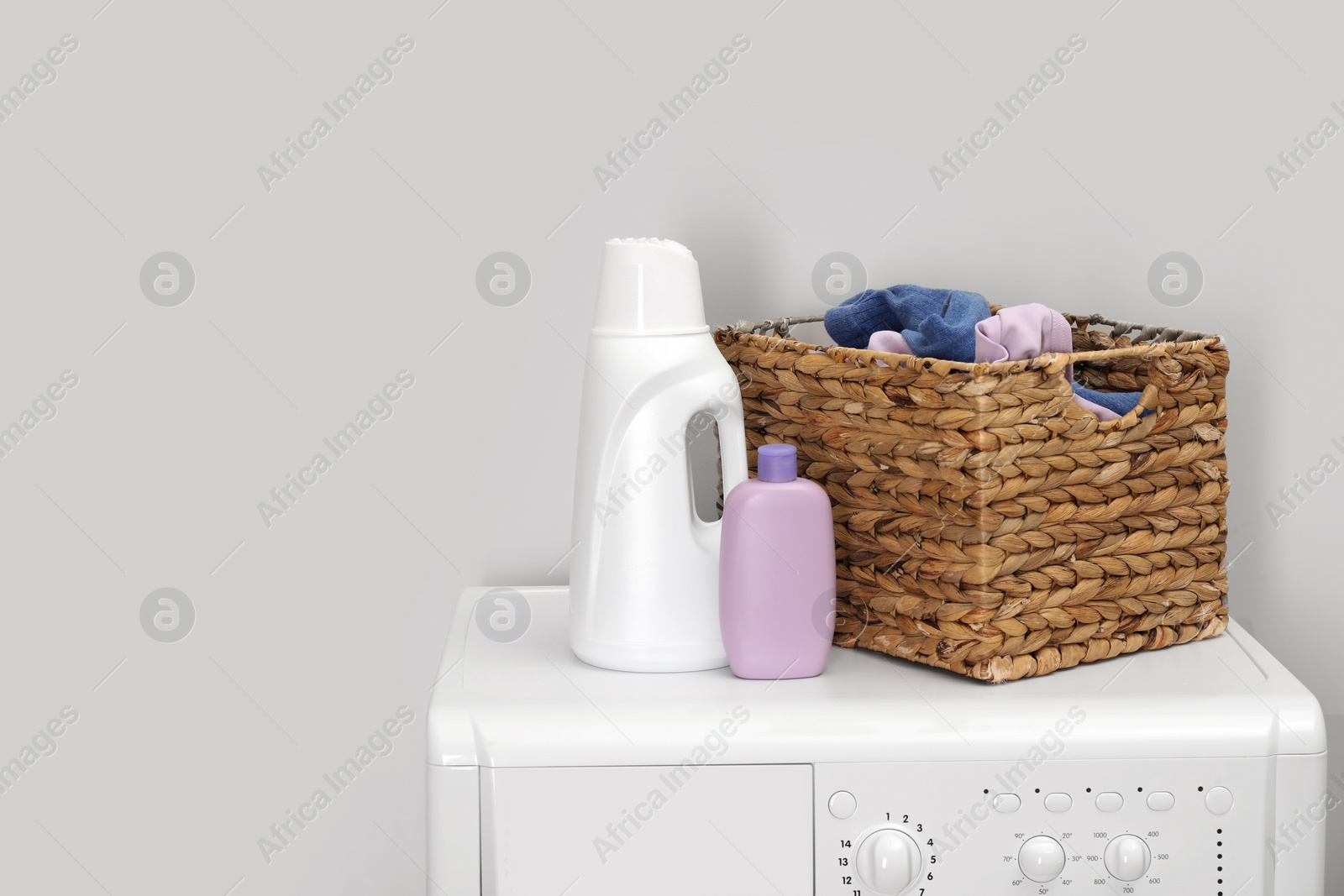 Photo of Baby clothes in wicker basket and laundry detergents on washing machine near light wall, space for text