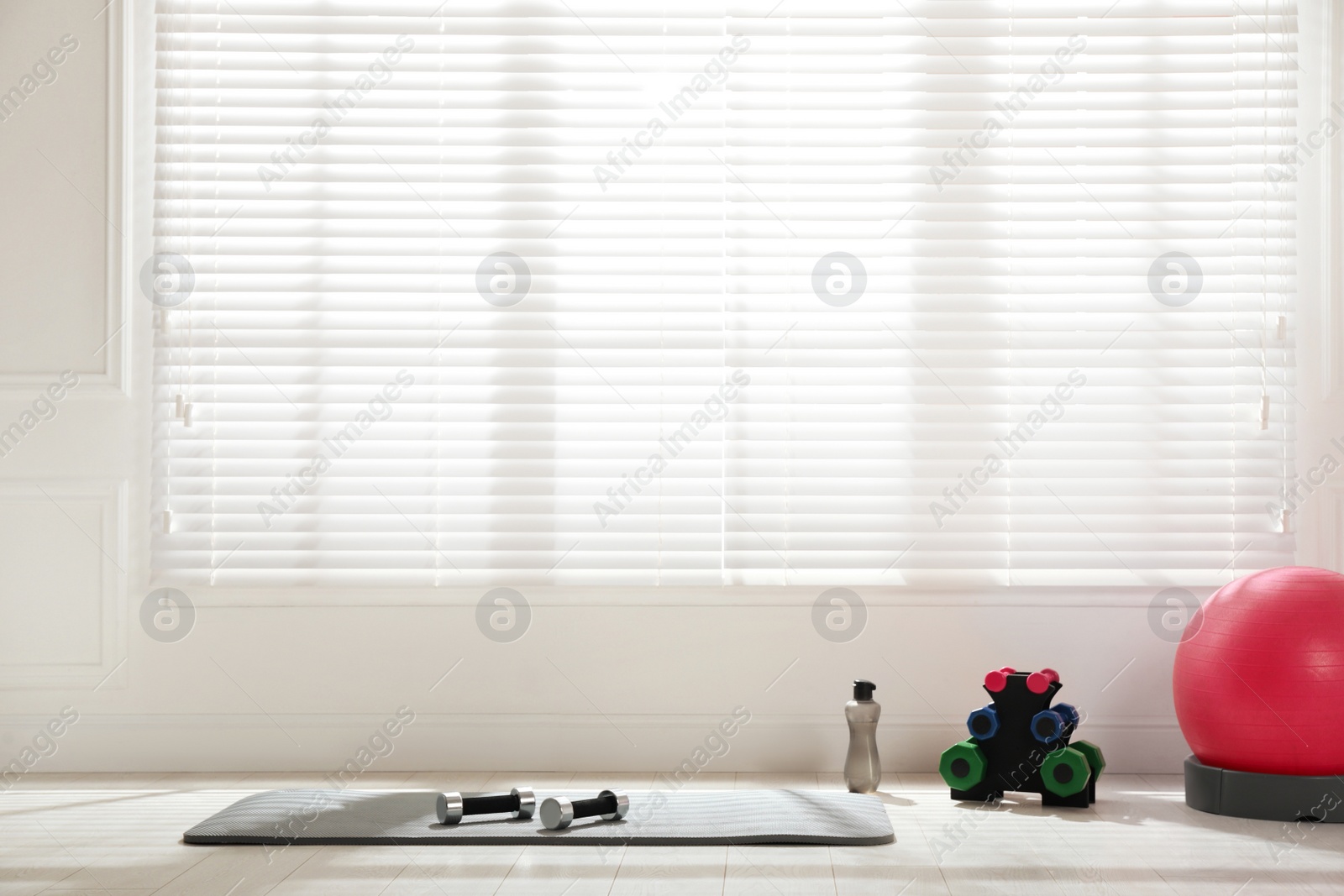 Photo of Exercise mat, dumbbells, fitness ball and bottle of water near window in spacious room