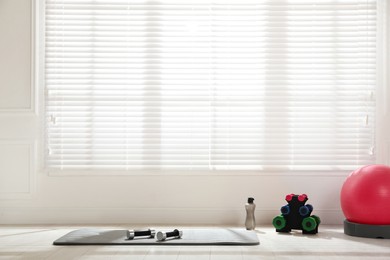 Photo of Exercise mat, dumbbells, fitness ball and bottle of water near window in spacious room