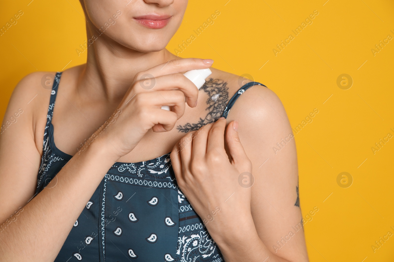 Photo of Woman applying cream onto tattoo on her skin against yellow background, closeup