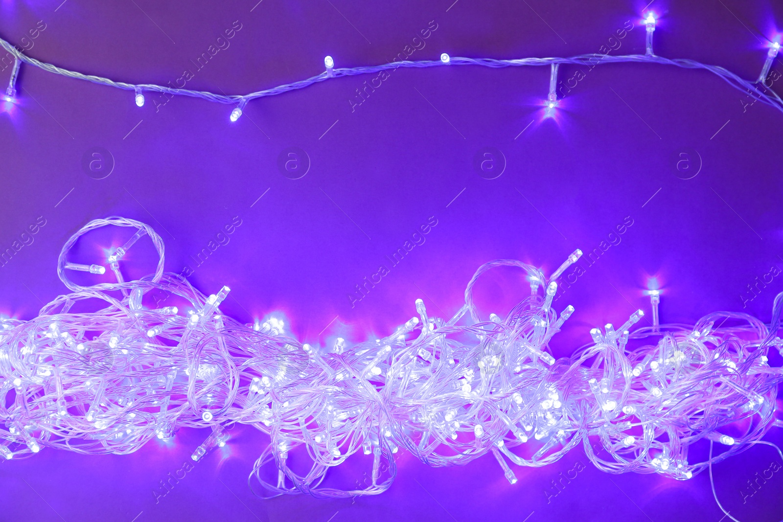 Photo of Glowing Christmas lights on violet  background, top view