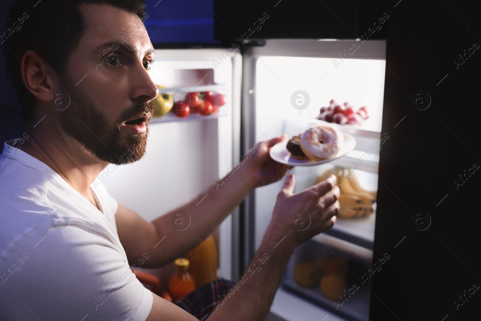 Photo of Man taking plate with donuts from refrigerator in kitchen at night. Bad habit