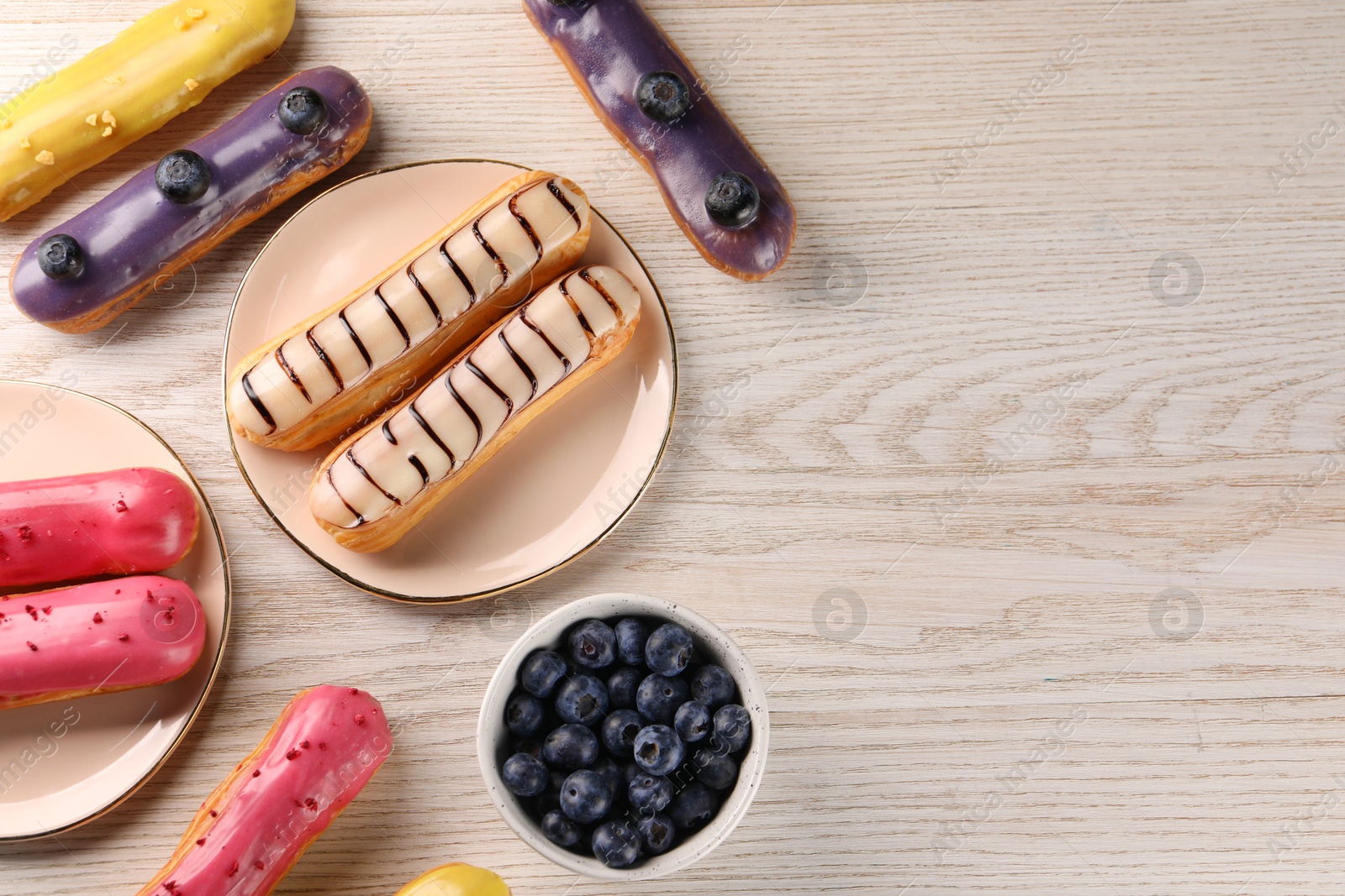 Photo of Different tasty glazed eclairs and blueberries on light wooden table, flat lay. Space for text