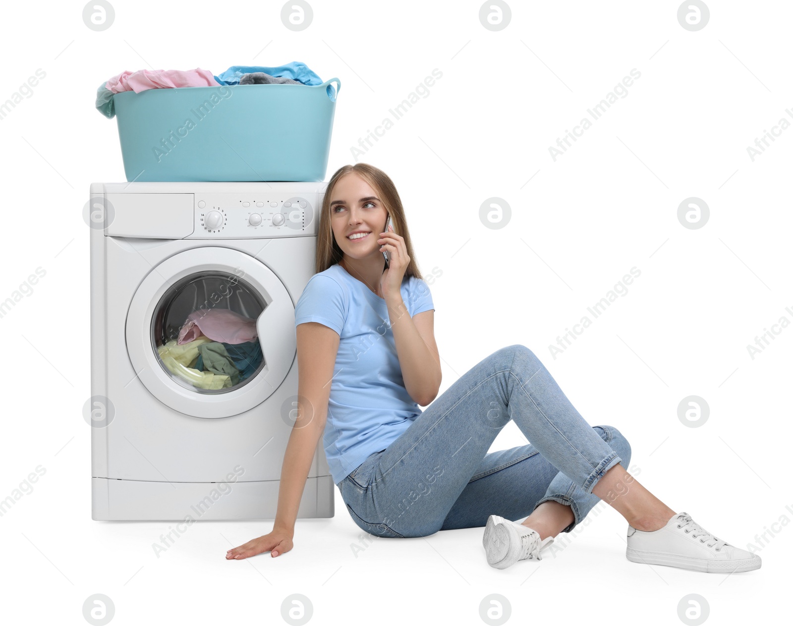 Photo of Beautiful young woman talking on smartphone near washing machine with laundry against white background