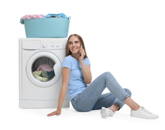 Beautiful young woman talking on smartphone near washing machine with laundry against white background