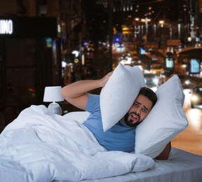 Image of Man covering ears with pillows in bed and beautiful view of night cityscape on background. Poor sleep because of urban noise