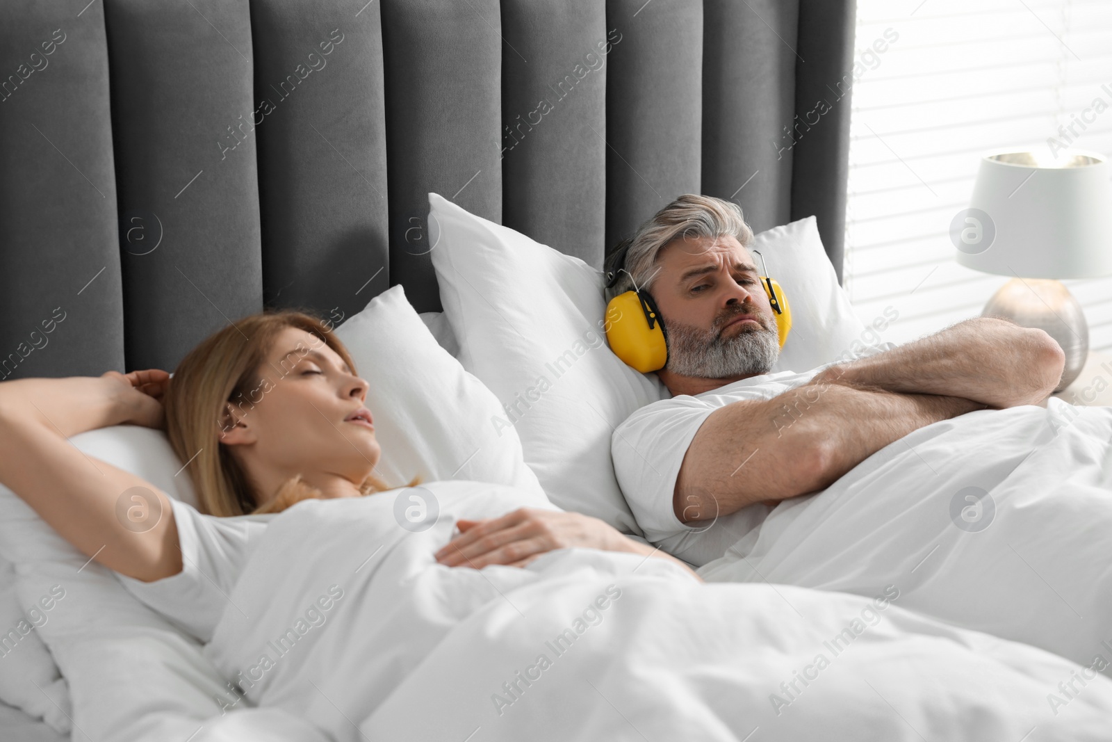 Photo of Irritated man with headphones lying near his snoring wife in bed at home