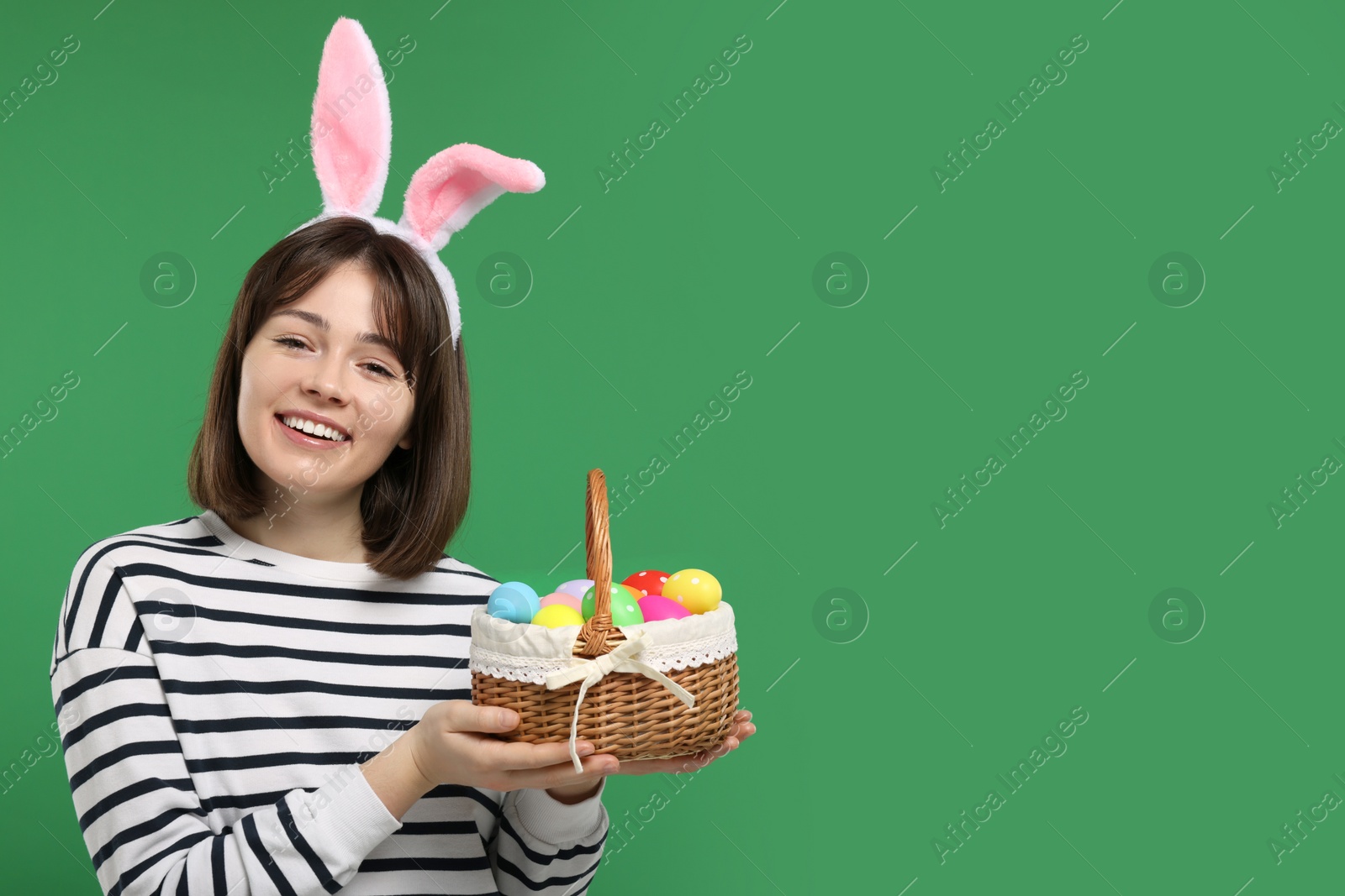 Photo of Easter celebration. Happy woman with bunny ears and wicker basket full of painted eggs on green background, space for text