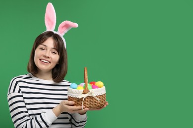 Easter celebration. Happy woman with bunny ears and wicker basket full of painted eggs on green background, space for text