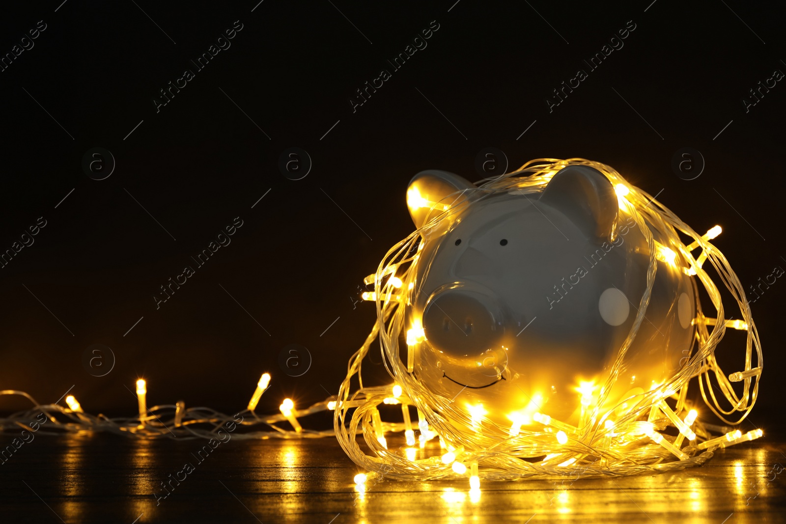 Photo of Piggy bank with Christmas lights on wooden table against black background. Space for text