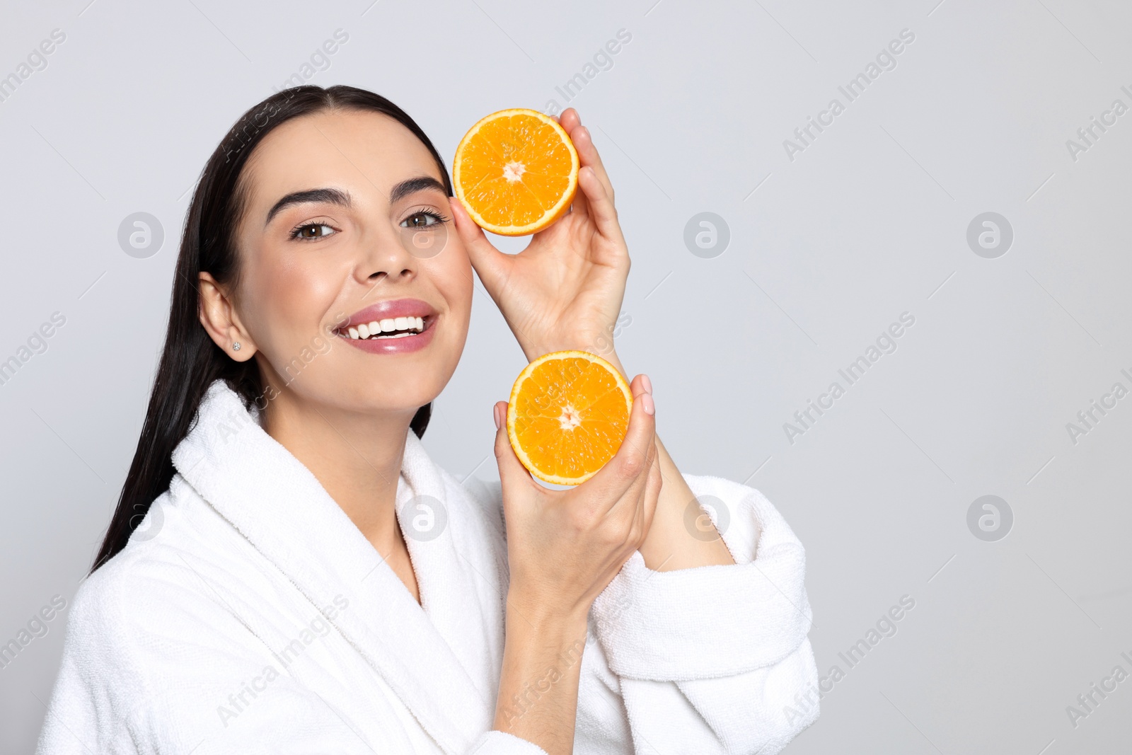 Photo of Woman in bathrobe holding juicy cut orange on light grey background, space for text. Spa treatment