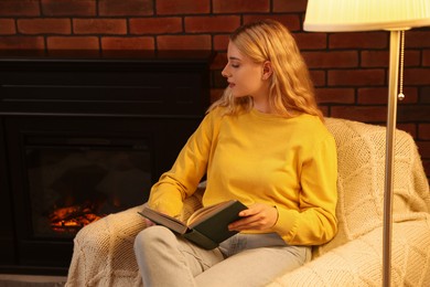 Photo of Beautiful young woman reading book near fireplace in room